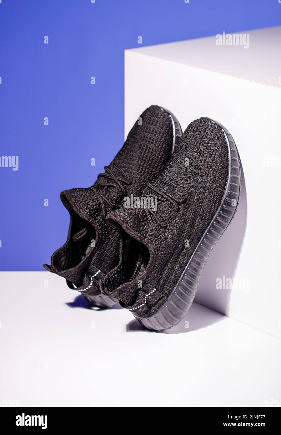 Schwarze Sneakers auf blauem Hintergrund mit weißem Podium in Form eines geometrischen Würfels. Seitenansicht der Turnschuhe im Unisex-Stil, die auf dem Podium stehen. Feder A Stockfoto