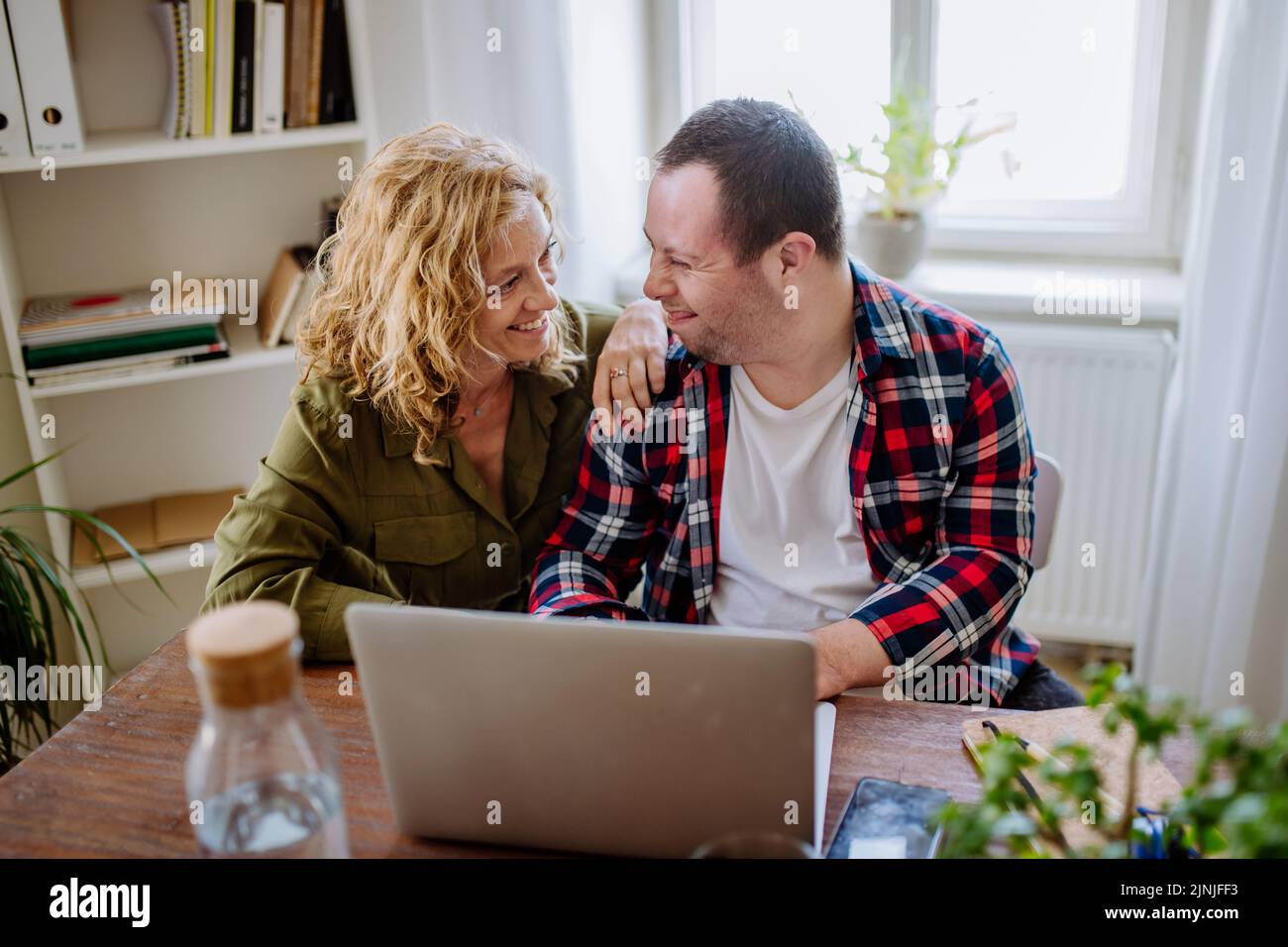 Der junge Mann mit Down-Syndrom sitzt am Schreibtisch im Büro und benutzt einen Laptop, seine Mutter hilft ihm. Stockfoto