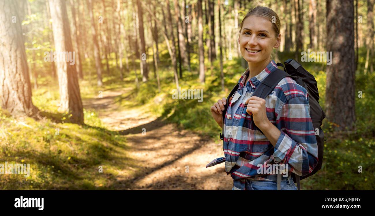 Lächelnde junge Frau im karierten Hemd mit Rucksack auf Waldweg stehen. Natur Abenteuer Wanderung. Copy space Stockfoto