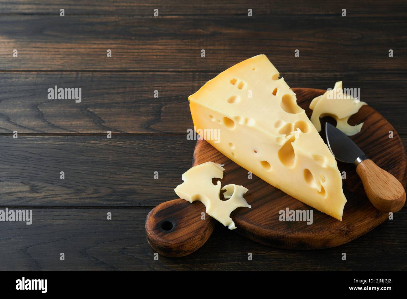 Käse. Stück Käsedreieck, Schweizer Käse mit Basilikum und ein Messer auf einem Servierbrett auf dunklem Holztischhintergrund. Käsehintergrund. Stockfoto