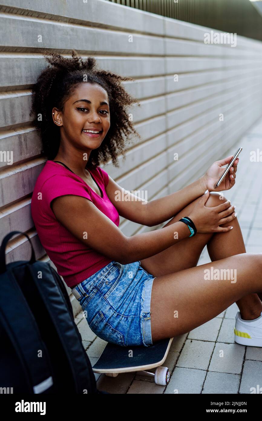 Multirassisches Teenager-Mädchen mit Rucksack, das am Skateboard sitzt und während des Sommers das Telefon benutzt. Stockfoto