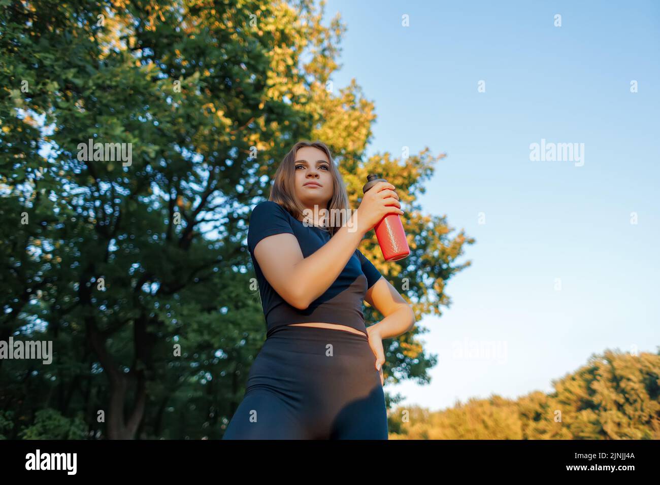 Die Fitness-blonde Frau in einem schwarzen Trainingsanzug hält eine rote Flasche Wasser in den Händen und löscht ihren Durst nach einem harten Training Stockfoto