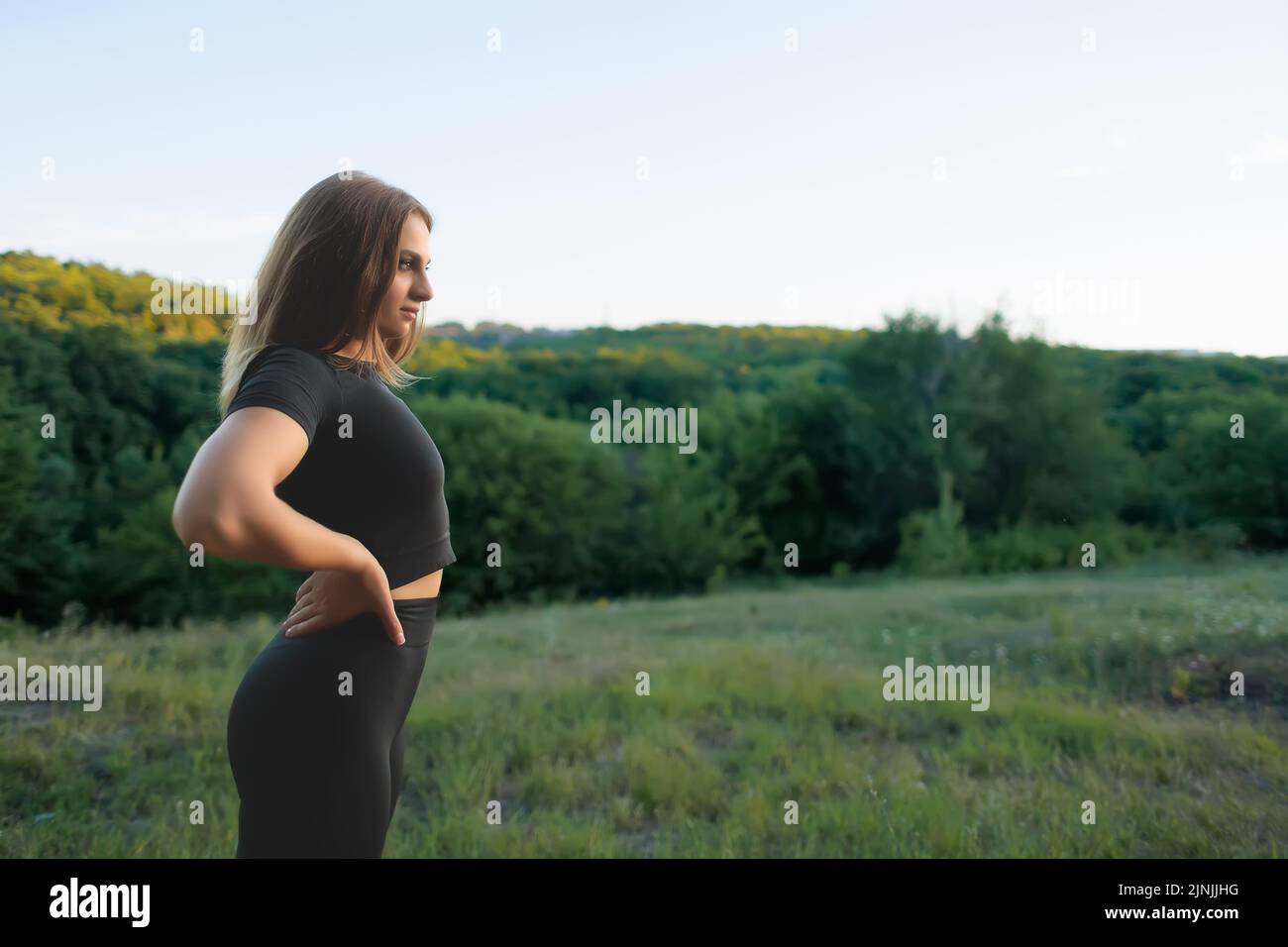 Eine junge Sportlerin in einem schwarzen Trainingsanzug hält ihre Hände am Gürtel und blickt weg auf den Sonnenuntergang im Park Stockfoto