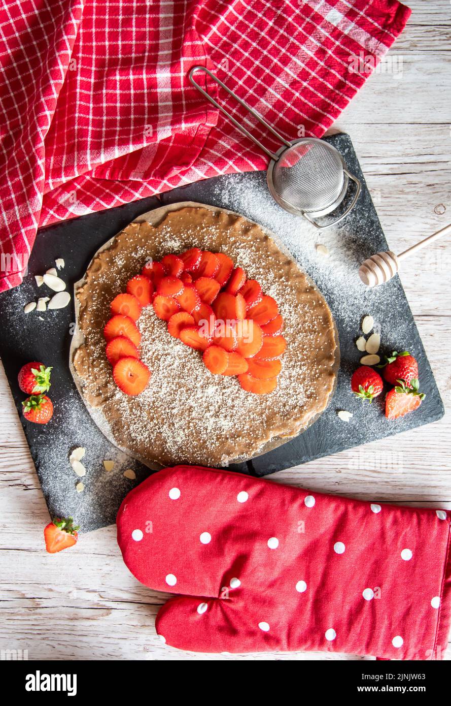 Hausgemachter Galette-Kuchen mit Erdbeeren und Mandel, auf schwarzem Schreibtisch und Holztisch platziert. Rotes Handtuch mit Kochwerkzeugen an der Seite Stockfoto