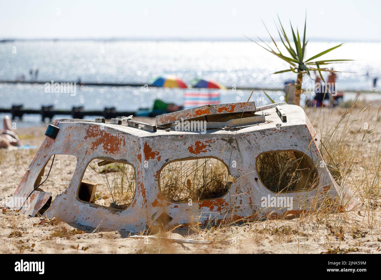 Ein kaputtes und verlassene Bootsdach an einem Sandstrand mit Gräsern, die innen und um sich wachsen und Menschen im Meer verschwimmen im Hintergrund Stockfoto