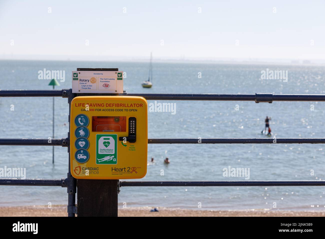 Ein öffentlicher lebensrettender Defibrillator in einem verschlossenen Kasten auf Metallgeländern mit Strand- und Meereshintergrund Stockfoto