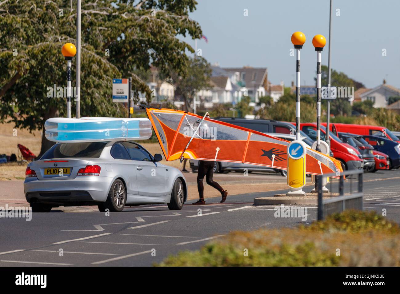 An einem sonnigen Tag wartet ein Auto auf einen Mann mit Windsurfsegel, der an einer Fußgängerüberfahrt die Straße überquert Stockfoto