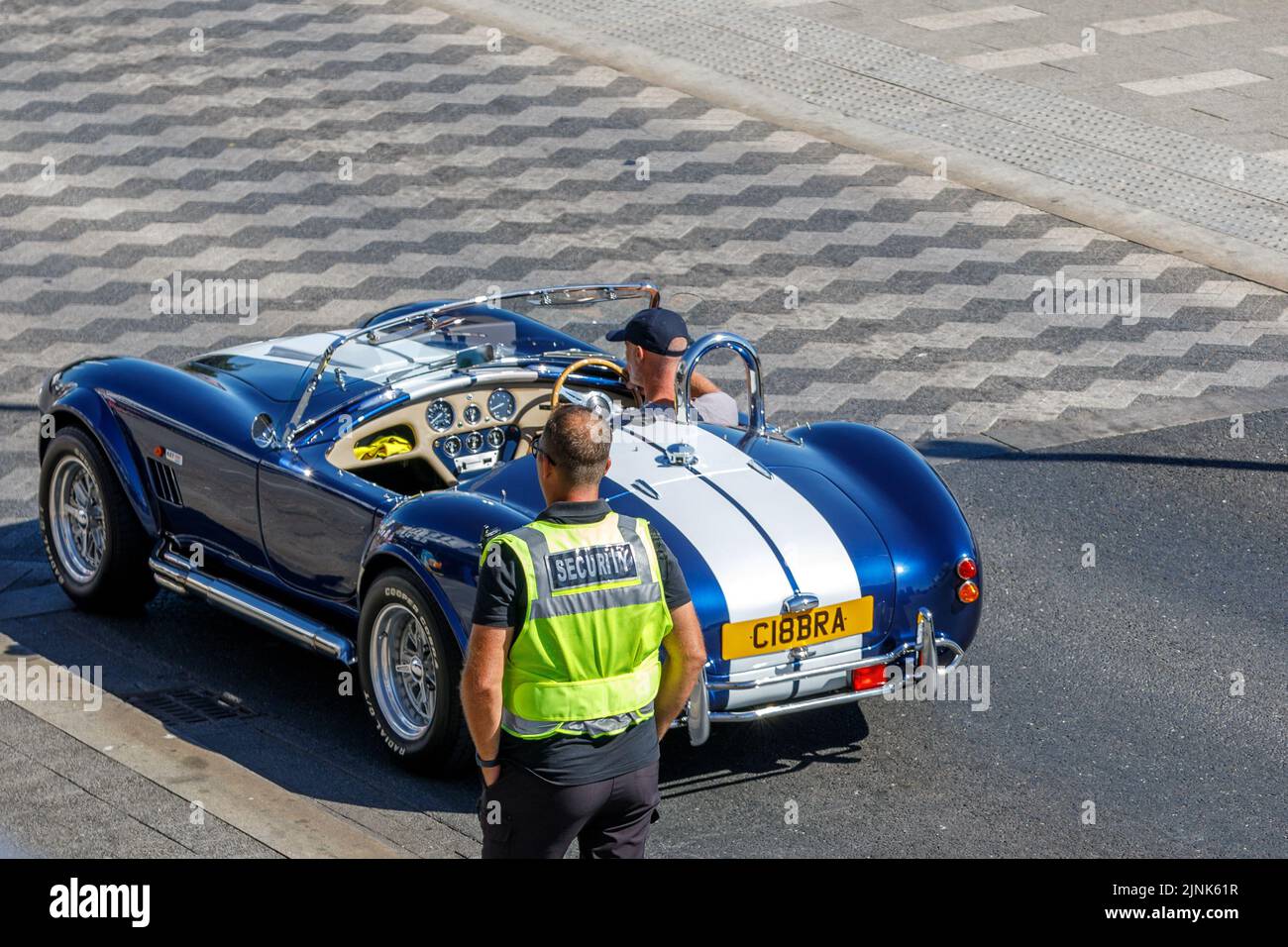 Ein Sicherheitsbeamter schaut auf ein blaues AC Cobra Auto mit weißem Streifen, das an einem heißen, sonnigen Tag entlang einer englischen Straße fährt, hohe Aussicht von oben Stockfoto