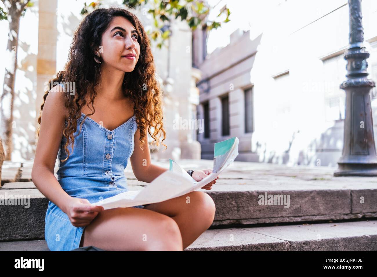 Hispanische Touristenin mit einem Stadtplan, der auf einer Treppe sitzt und tagsüber auf die sonnenbeschienenen Stadtstraßen blickt Stockfoto