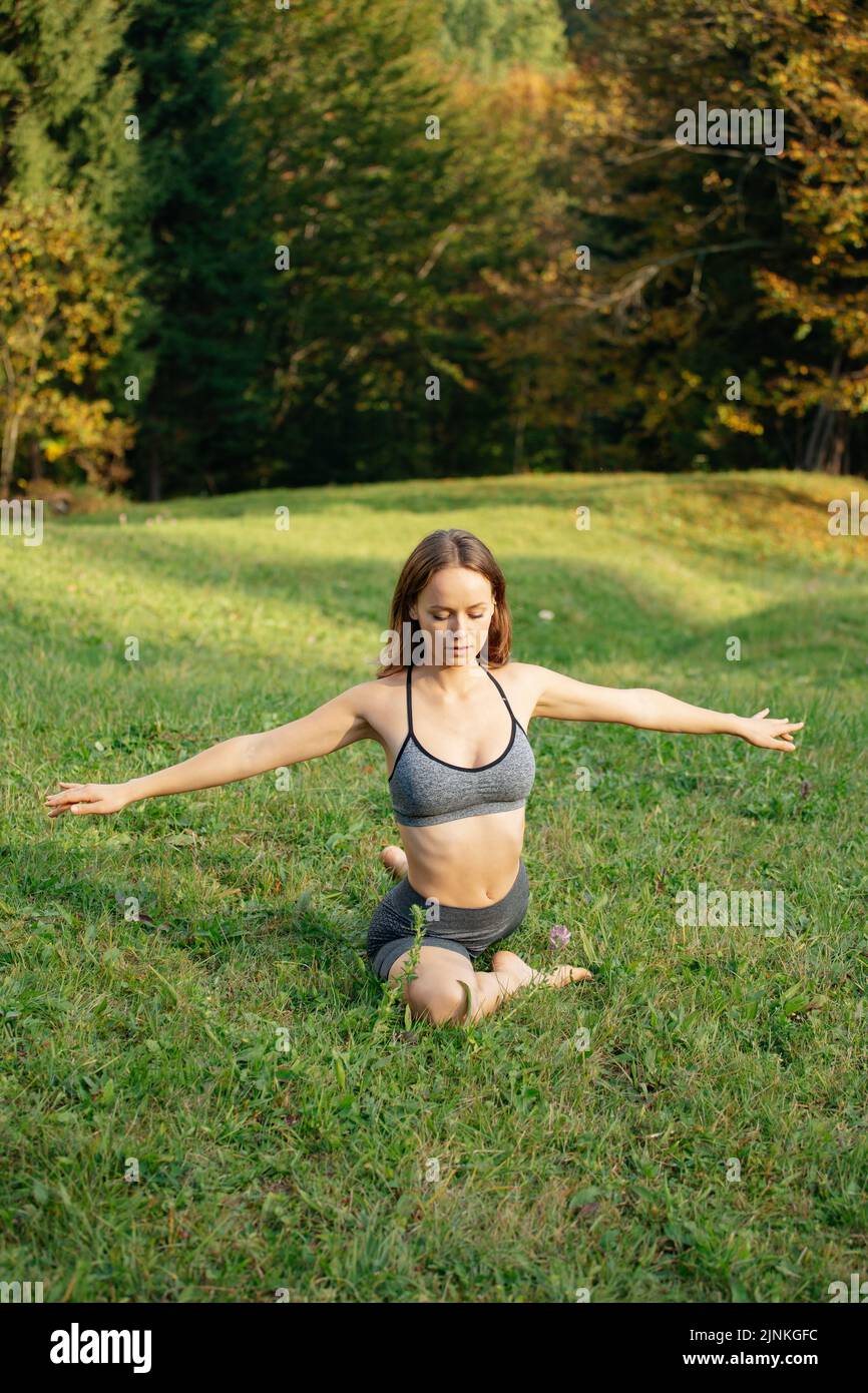 Seitenansicht einer sportlichen Frau mit ausgestreckten Armen beim Stretching auf Gras im herbstlichen Bergwald. Konzept des Outdoor-Trainings, Körper Stockfoto
