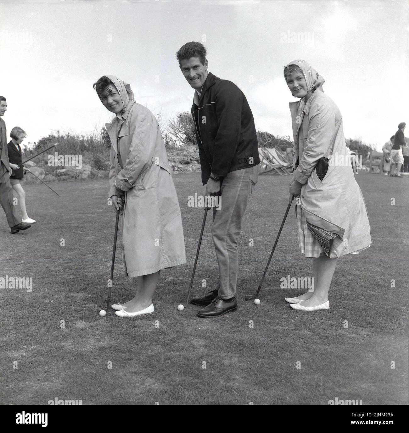 1950s, historisch, draußen, zwei junge Frauen in Mänteln und Kopftüchern, mit einem jungen Mann, der auf Gras steht, für ein Foto, mit Puttern, auf einem Golfplatz & Putt, Isle of Wright, England, Großbritannien. Stockfoto