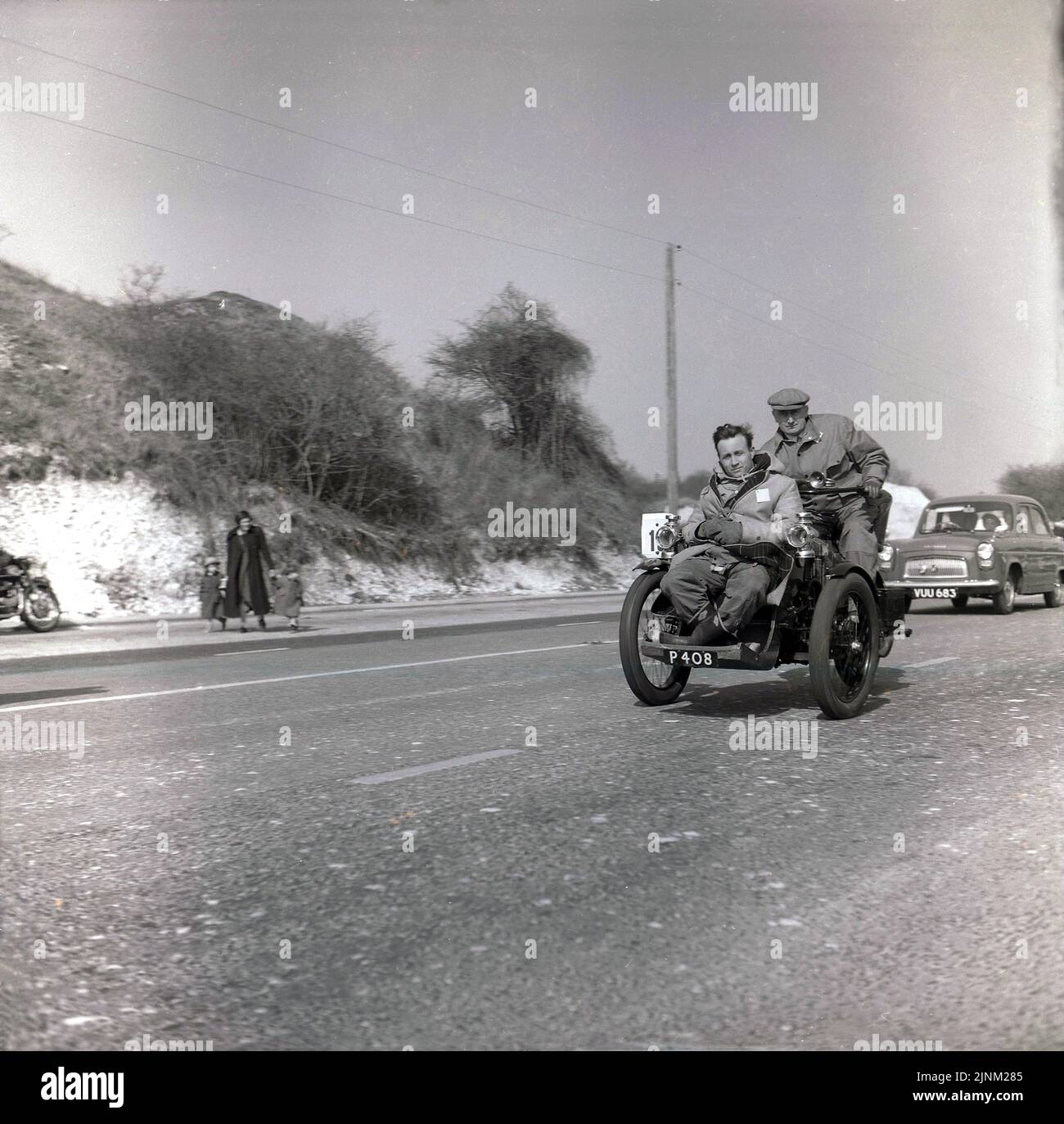 1960, historisch, London bis Brighton Veteran Car Run, auf der Brighion Road (A23) zwei Männer, die in einem oben offenen dreirädrigen motorisierten Fahrzeug, einem Motor-Dreirad (Kennzeichen P 408), fahren... hoffen, dass der vorne sitzende Herr in jenen Tagen Natürlich. in ...no Sturzhelme geschnallt wird Um am Oldtimerlauf teilzunehmen, müssen alle Fahrzeuge prre 1905 sein. Ein Ford 100E der Ära auf der Straße hinter gesehen. Stockfoto