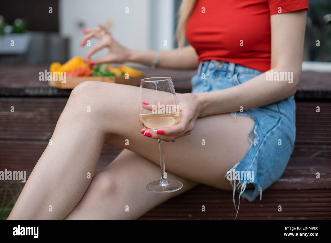 Zugeschnittenes Foto einer jungen Frau, die auf einer Stufe der hölzernen Veranda neben geschnittenen Früchten, Grüns sitzt und ein Glas Weißwein hält. Stockfoto