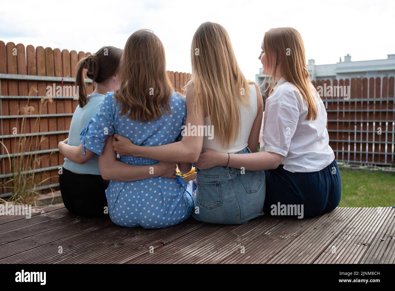 Rückansicht einer Gruppe junger Frauen mit langen Haaren, die auf einer hölzernen Veranda sitzen und sich im Hinterhof umarmen. Sommer. Stockfoto