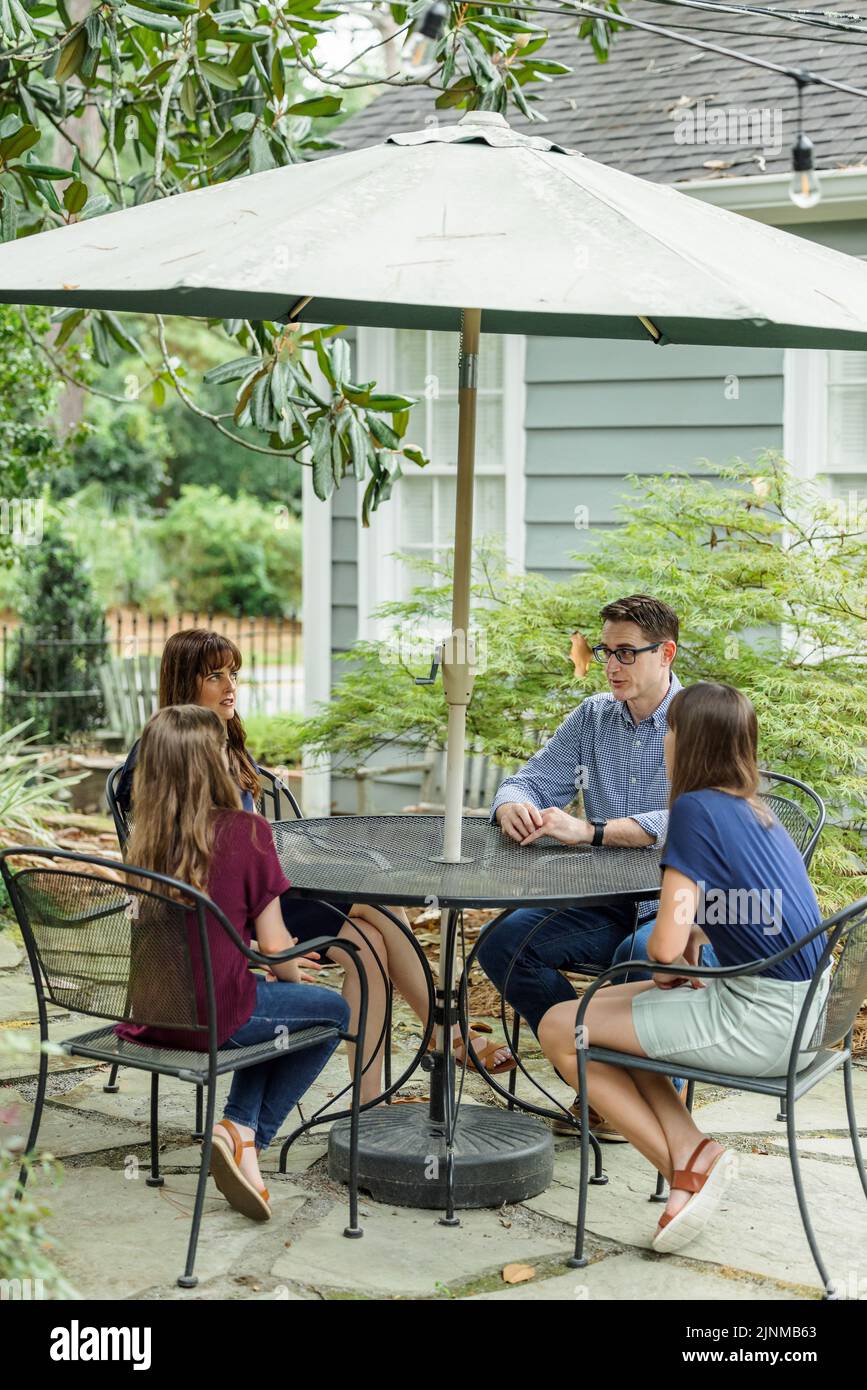 Eine Familie aus Mutter, Vater und zwei Töchtern, die draußen an einem Tisch auf der Terrasse sitzen und einen Regenschirm über ihren Tag reden. Stockfoto