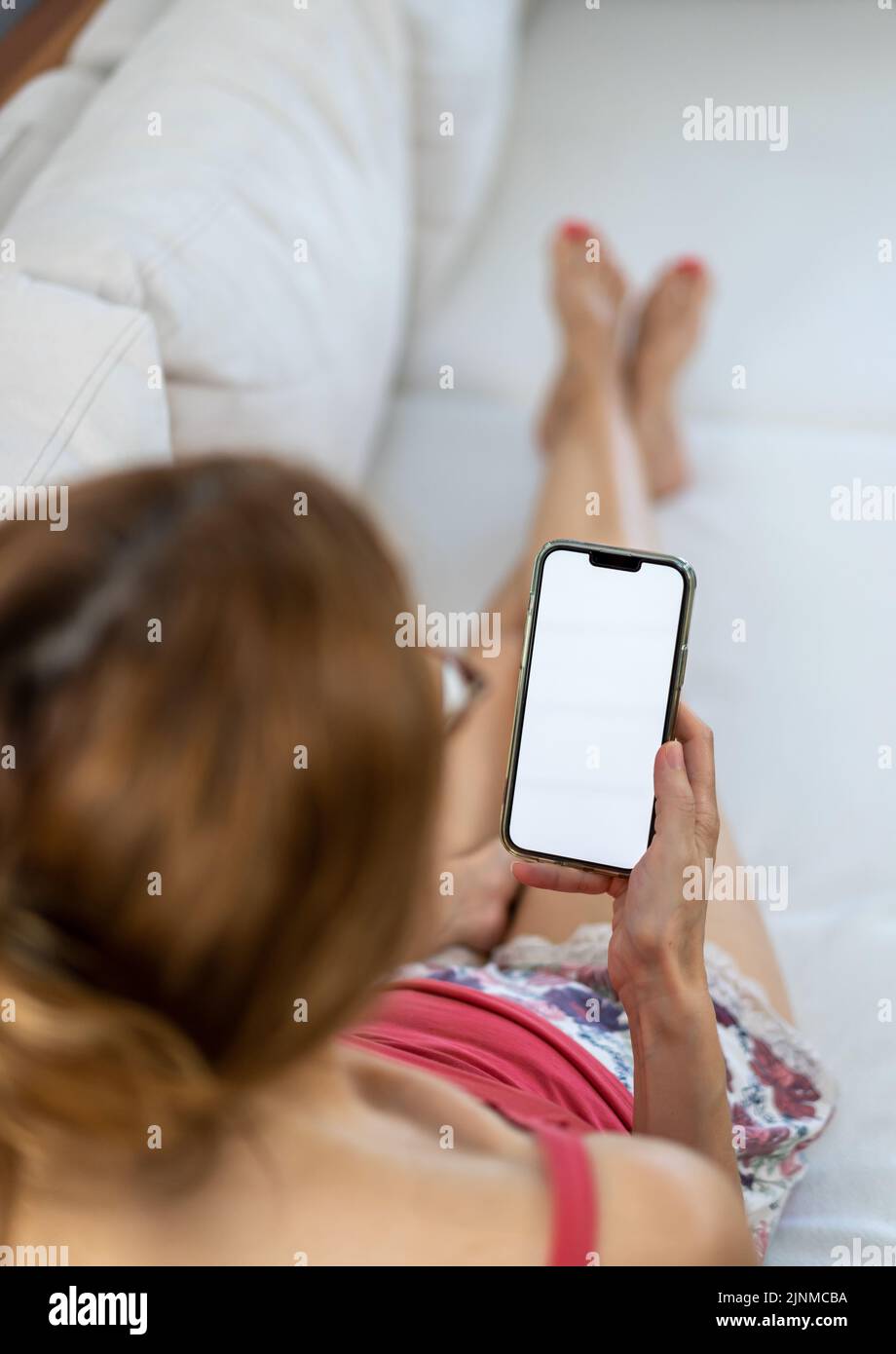 Frau, die auf dem weißen Sofa im Wohnzimmer liegt und zu Hause mit dem Mobiltelefon telefoniert, Vertikal Stockfoto