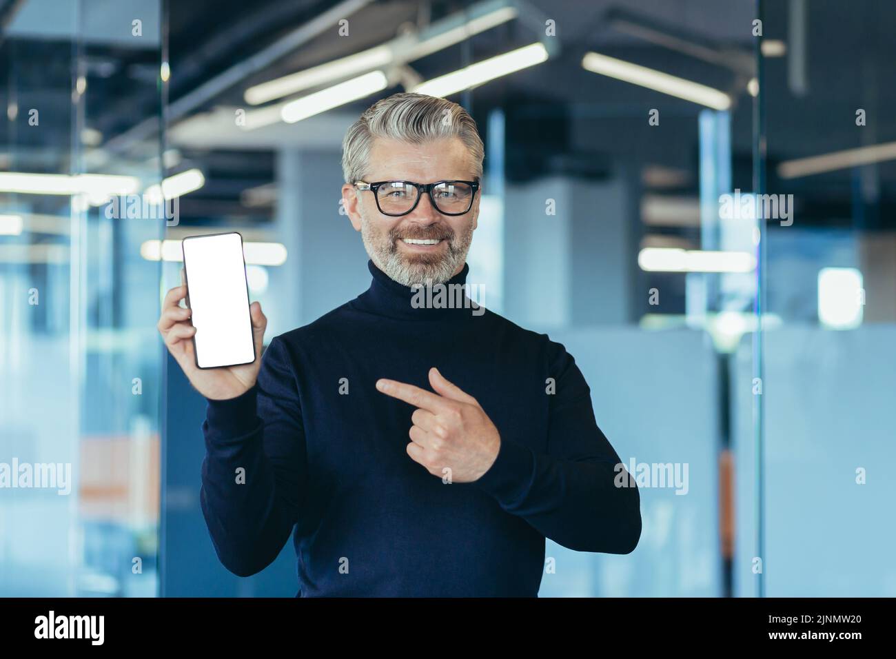 Leitender erfolgreicher grauhaariger Geschäftsmann im Büro, der der Kamera einen Telefonbildschirm mit weißem Hintergrund zeigt und mit dem Finger auf den Kopierbereich zeigt Stockfoto