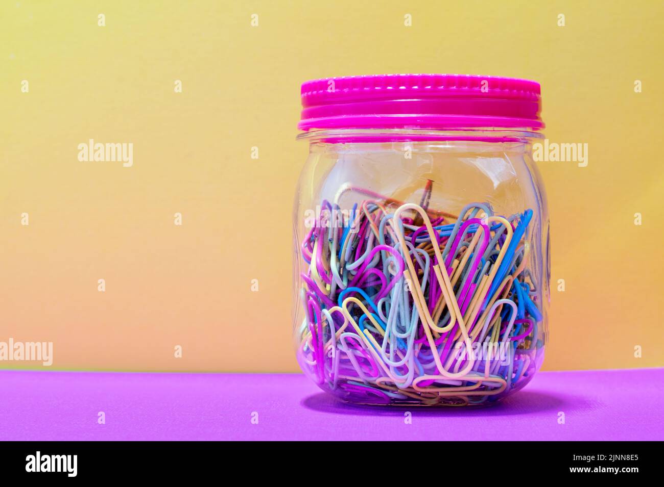Bunte Büroklammern in einem einfarbigen Glas und versiegeltem Deckel mit gelbem Hintergrund und Kopierraum, kreatives Konzept. Stockfoto