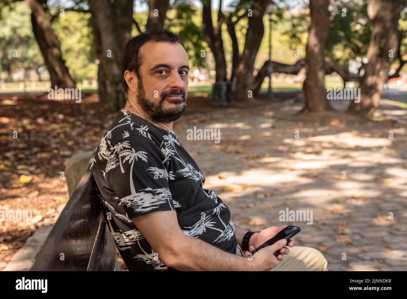 Ein Mann, der mit seinem Handy in der Hand auf einer Bank im Park sitzt und auf die Kamera schaut Stockfoto