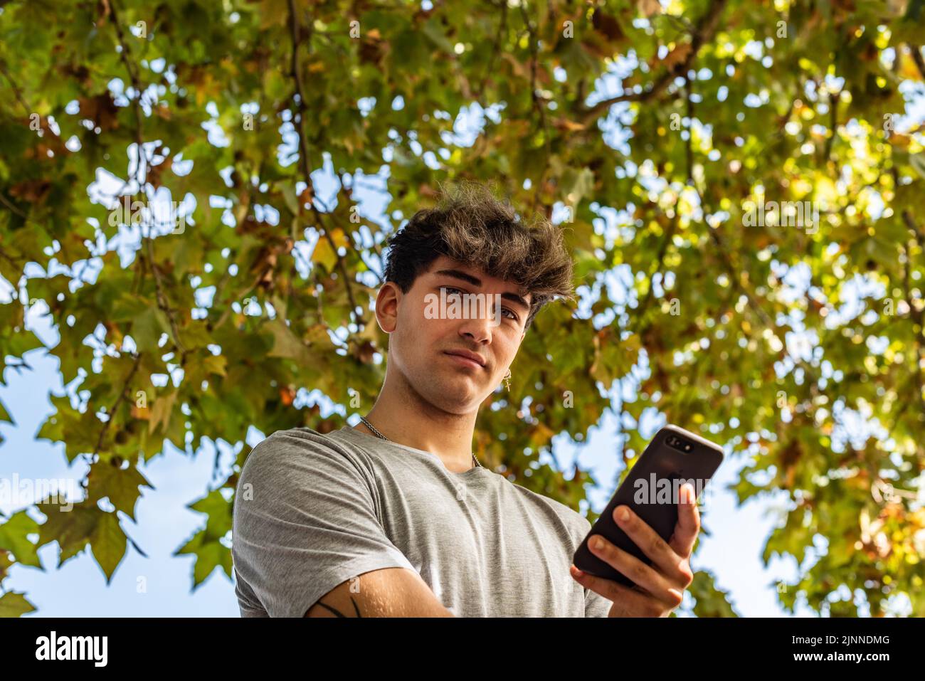 Niedriger Winkel eines jungen Mannes, der sein Smartphone in der Hand hält, ernst, und die Kamera in einem öffentlichen Park anschaut Stockfoto