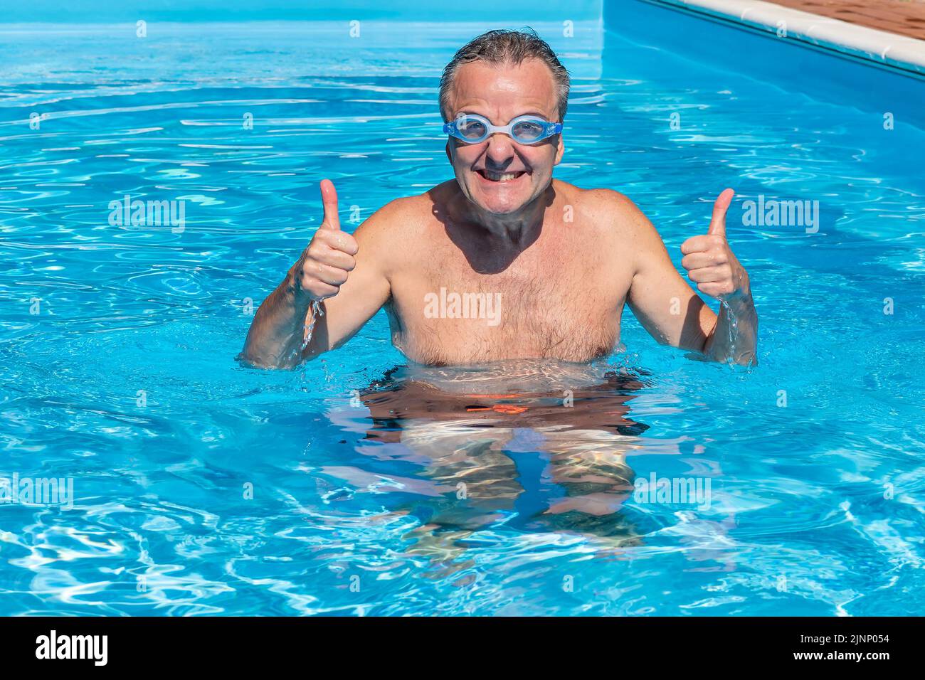 Ein weißer Mann mittleren Alters mit Daumen nach oben im Wasser eines Schwimmbades, mit einem sehr lustigen Ausdruck und einer Schwimmbrille Stockfoto