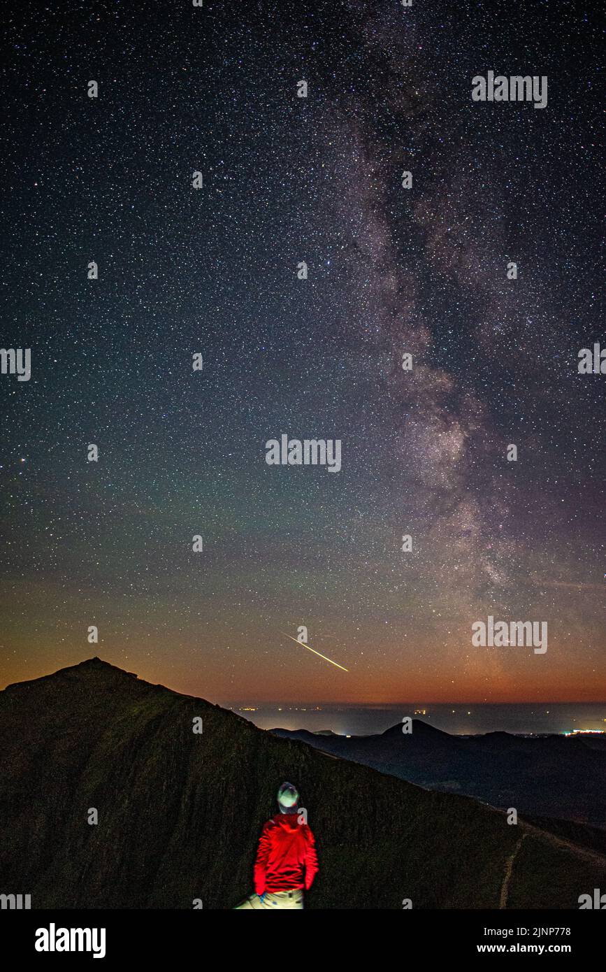Mann (Sterngucker), der den klaren Sternenhimmel der Nacht, die Milchstraße und den Feuerball, den Sternschnuppen (Meteor über dem Snowdon-Gipfel im Snowdonia-Nationalpark) beobachtet Stockfoto
