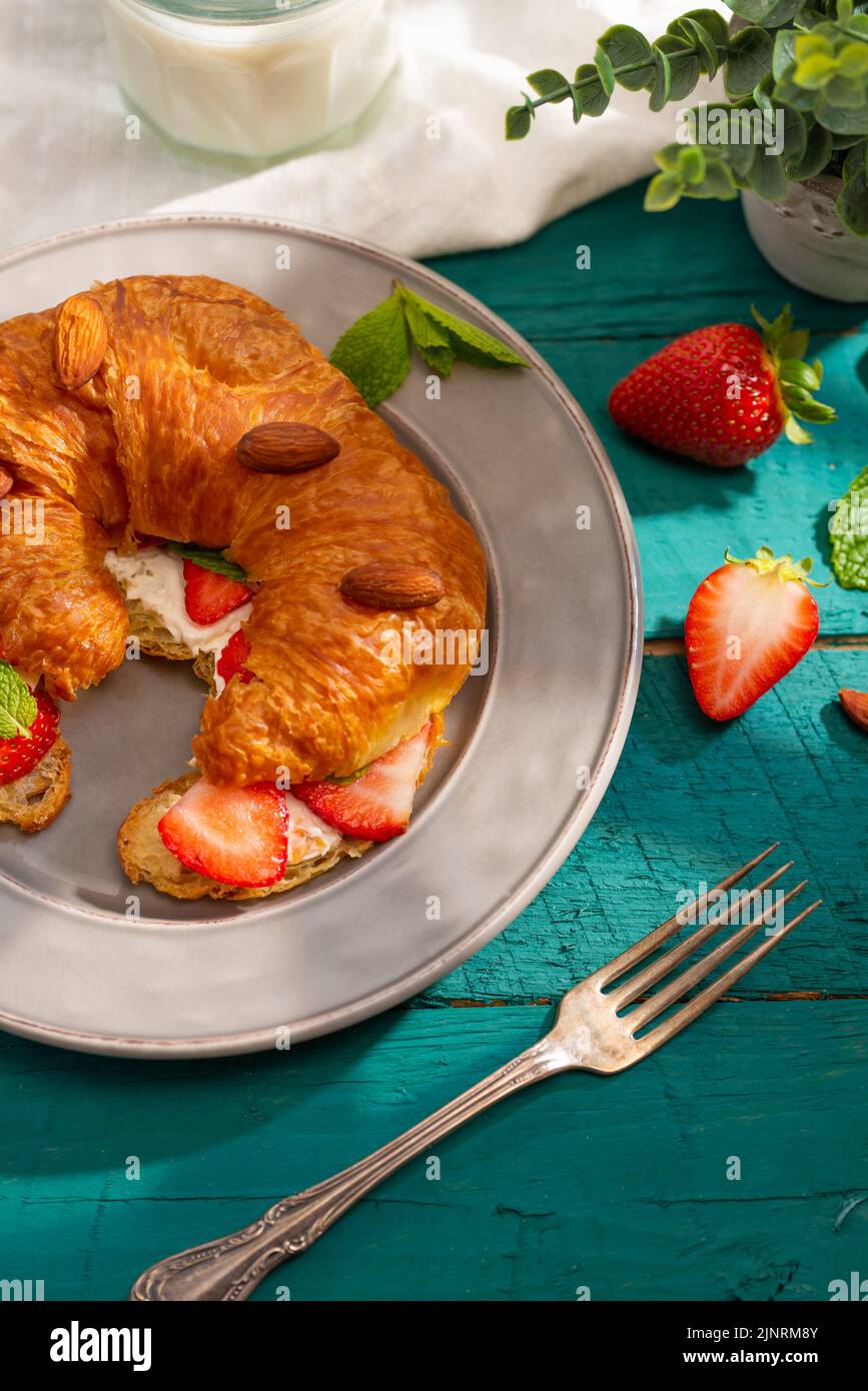 Appetitliches französisches Croissant mit Erdbeeren, Mandeln und duftenden Minzblättern auf einem Teller auf türkisfarbenem Hintergrund. Kontinentales Frühstück für eine person Stockfoto
