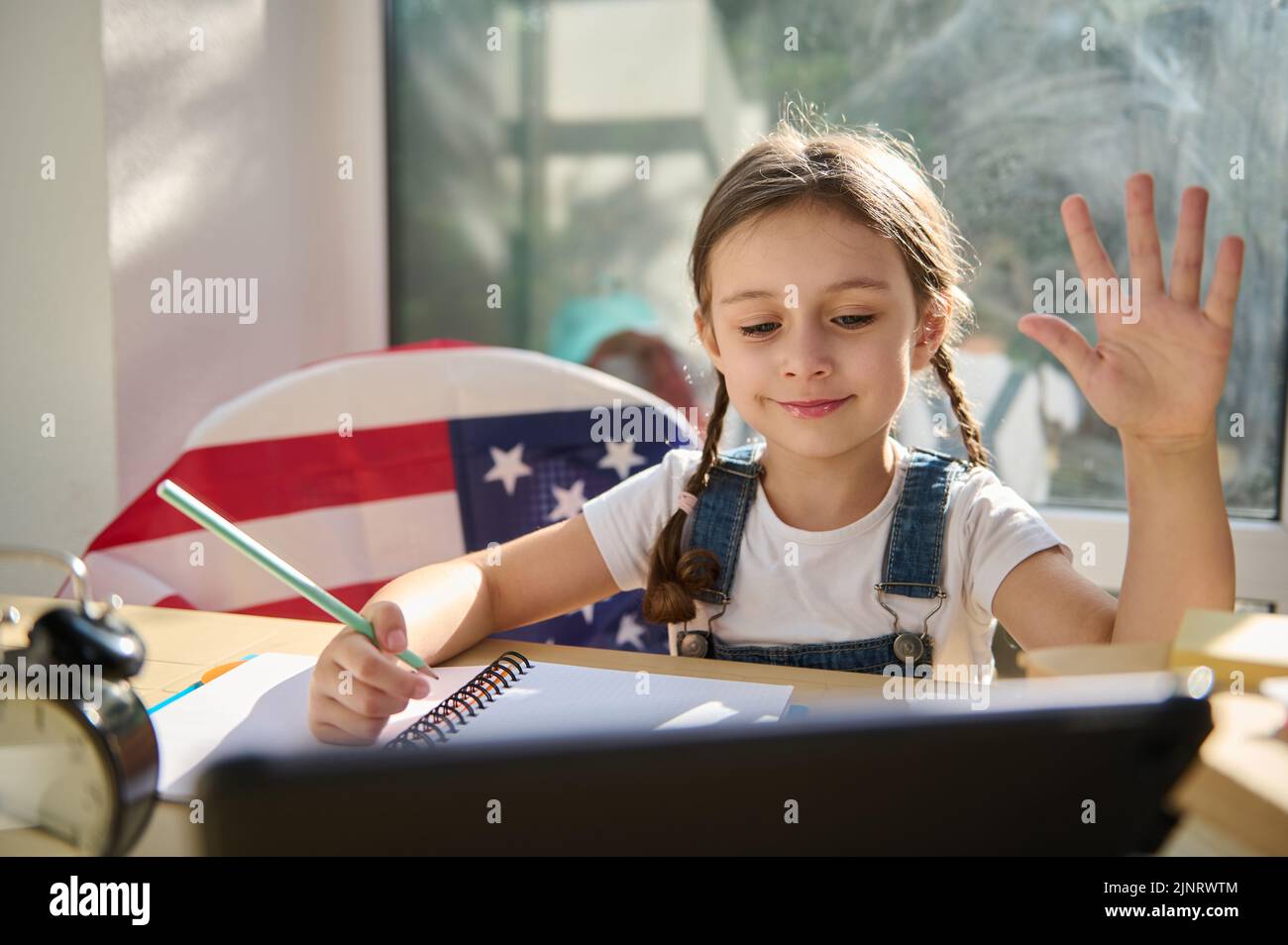 Schönes Kind, eine First-Grader winkt der Lehrerin die Hand zu, während sie sich eine Videostunde auf dem digitalen Tablet ansieht Stockfoto