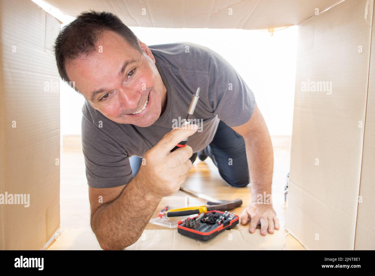 Fröhlicher Handwerker mit Werkzeug klettert in einen offenen Karton Stockfoto