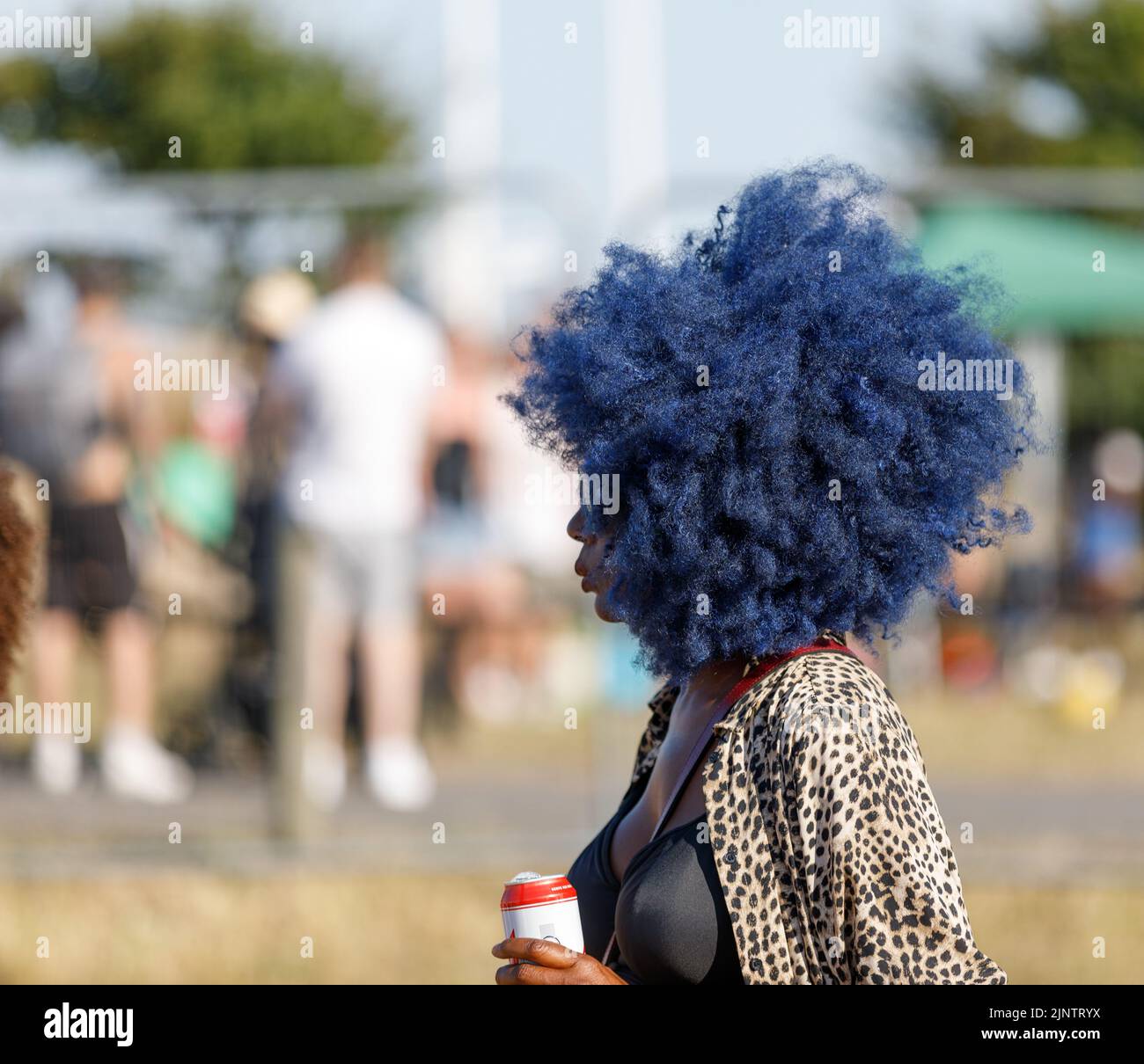 Eine schwarze Frau mit blauem Afro-Haar, eine grüne Sonnenbrille mit einer Bierkutte, draußen bei einem Festival, mit Menschen im Hintergrund, die nicht fokussiert sind Stockfoto