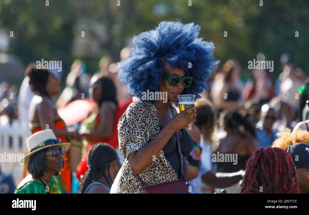 Eine schöne schwarze Frau mit blauem Afro-Haar, grüne Sonnenbrille, die draußen ein Bier trinkt und viele Menschen im Hintergrund außer Fokus stehen Stockfoto