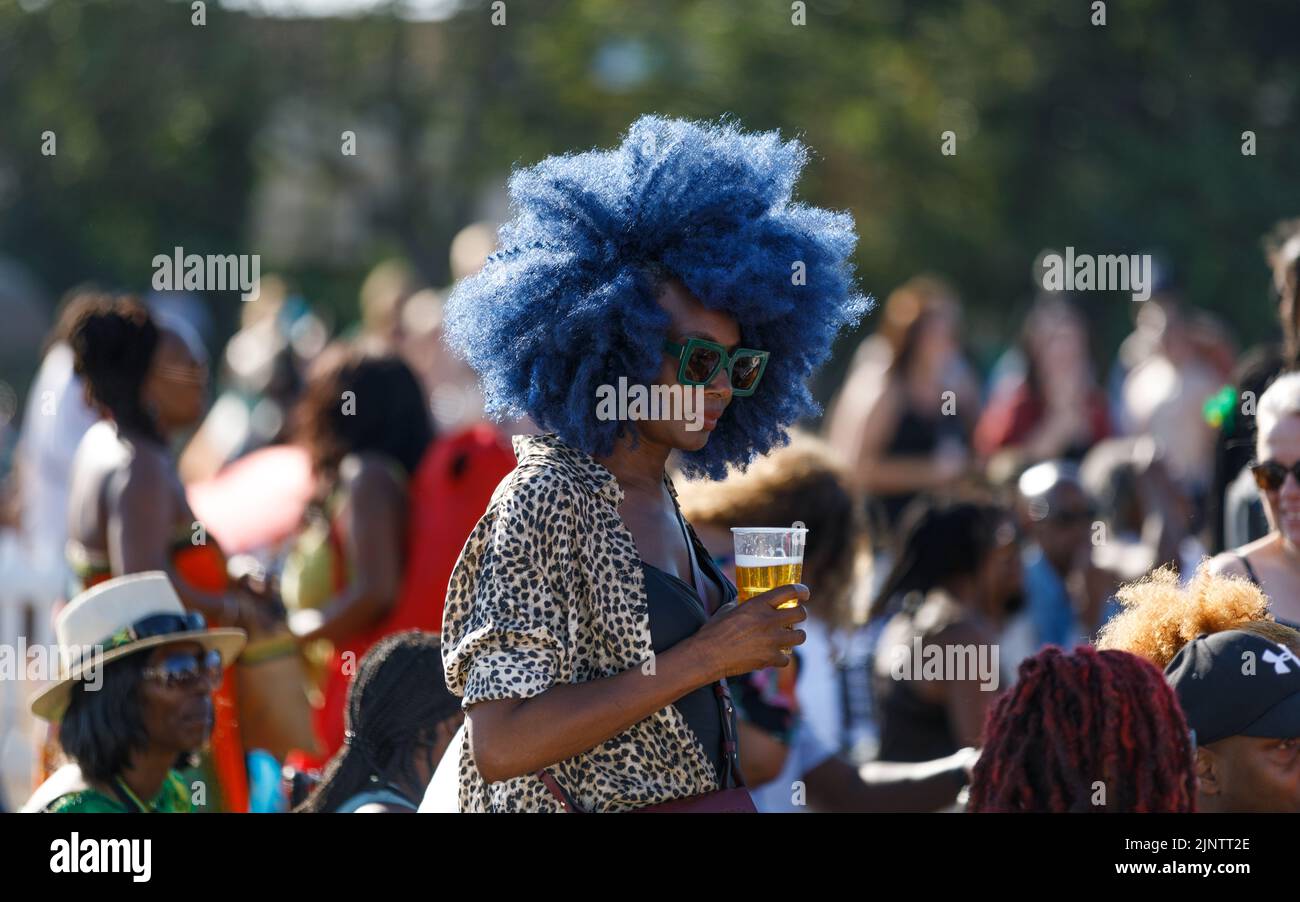 Eine schöne schwarze Frau mit blauem Afro-Haar, eine grüne Sonnenbrille, die ein Bierglas aus Kunststoff im Freien hält und viele Menschen im Hintergrund außer Fokus stehen Stockfoto