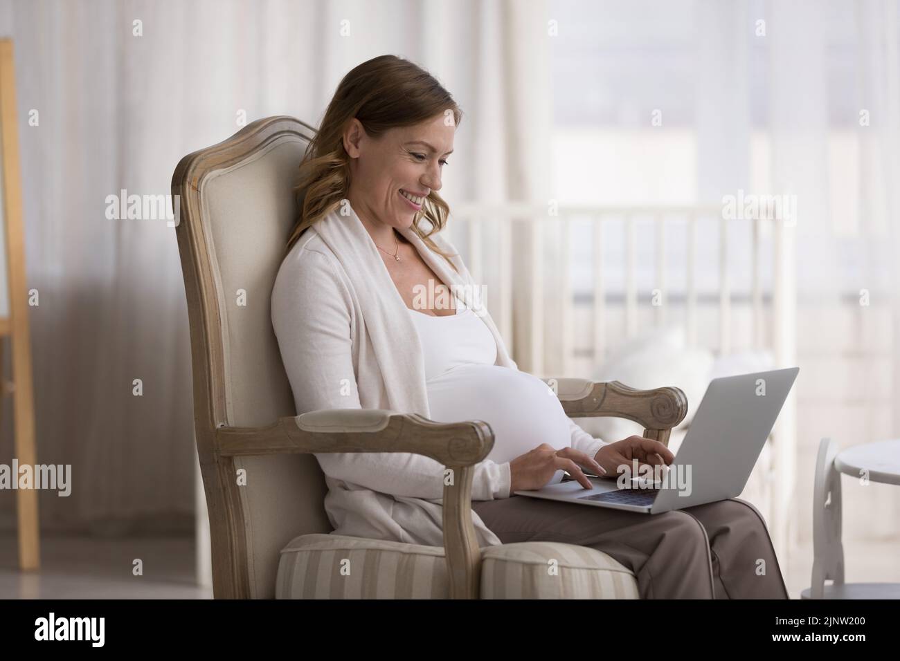 Junge, lächelnde Schwangerin entspannen sich auf einem Sessel mit Laptop Stockfoto