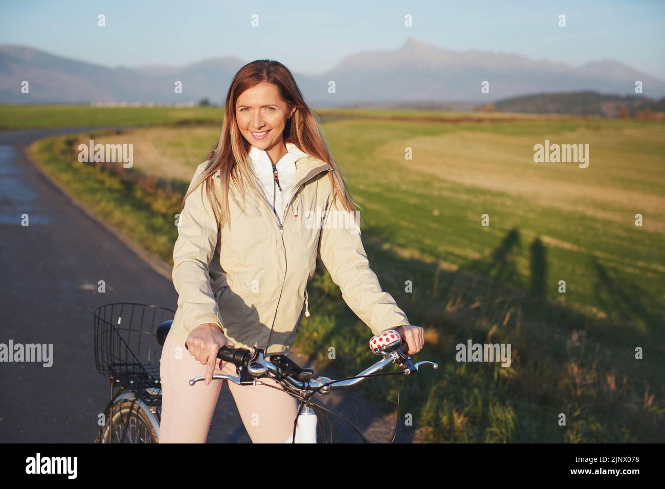 Junge Frau in Jacke steht mit dem Fahrrad auf Asphalt Landstraße, Nachmittag Sonne scheint auf Feldern Wiesen und Berge Hintergrund Stockfoto