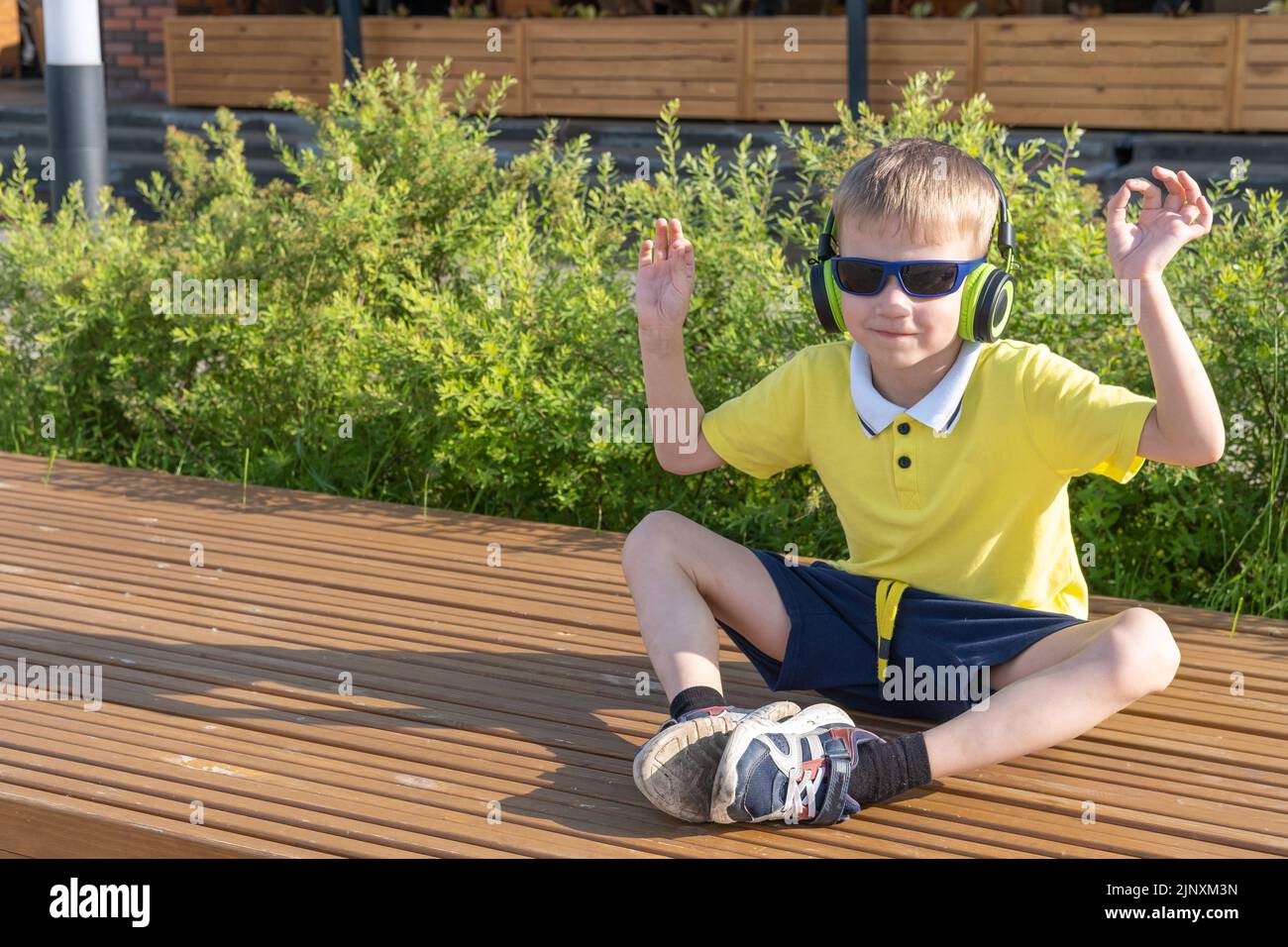 Ein stilvoller Junge mit Sonnenbrille und Kopfhörern hört seine Lieblingsmusik, während er auf einer Bank in einem Stadtpark sitzt. Ein glücklich lächelnder kleiner Junge genießt m Stockfoto