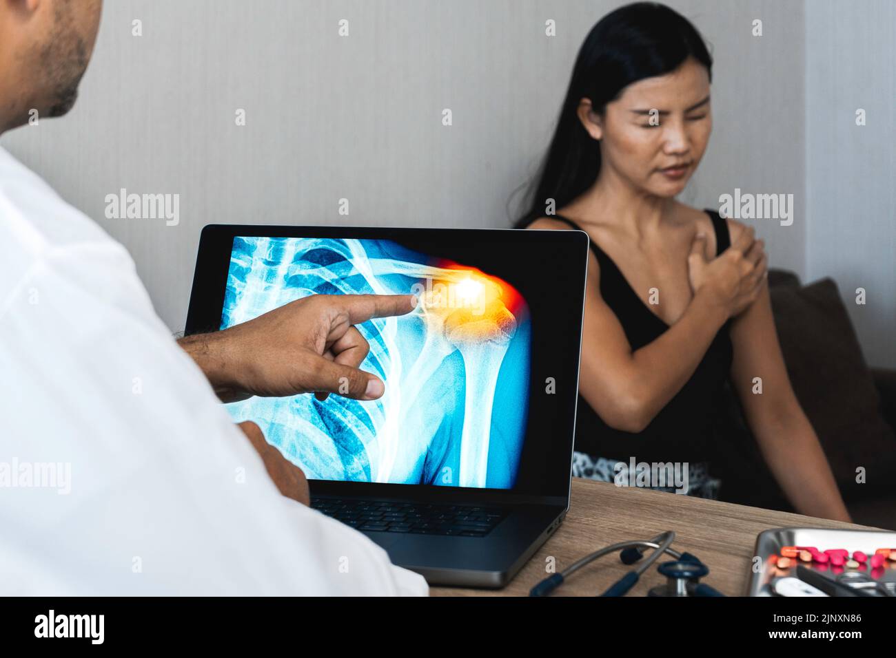 Arzt zeigt eine Röntgenaufnahme von Schmerzen in der Schulter auf einem Laptop. Patientin, die ihre Schulter hält Stockfoto