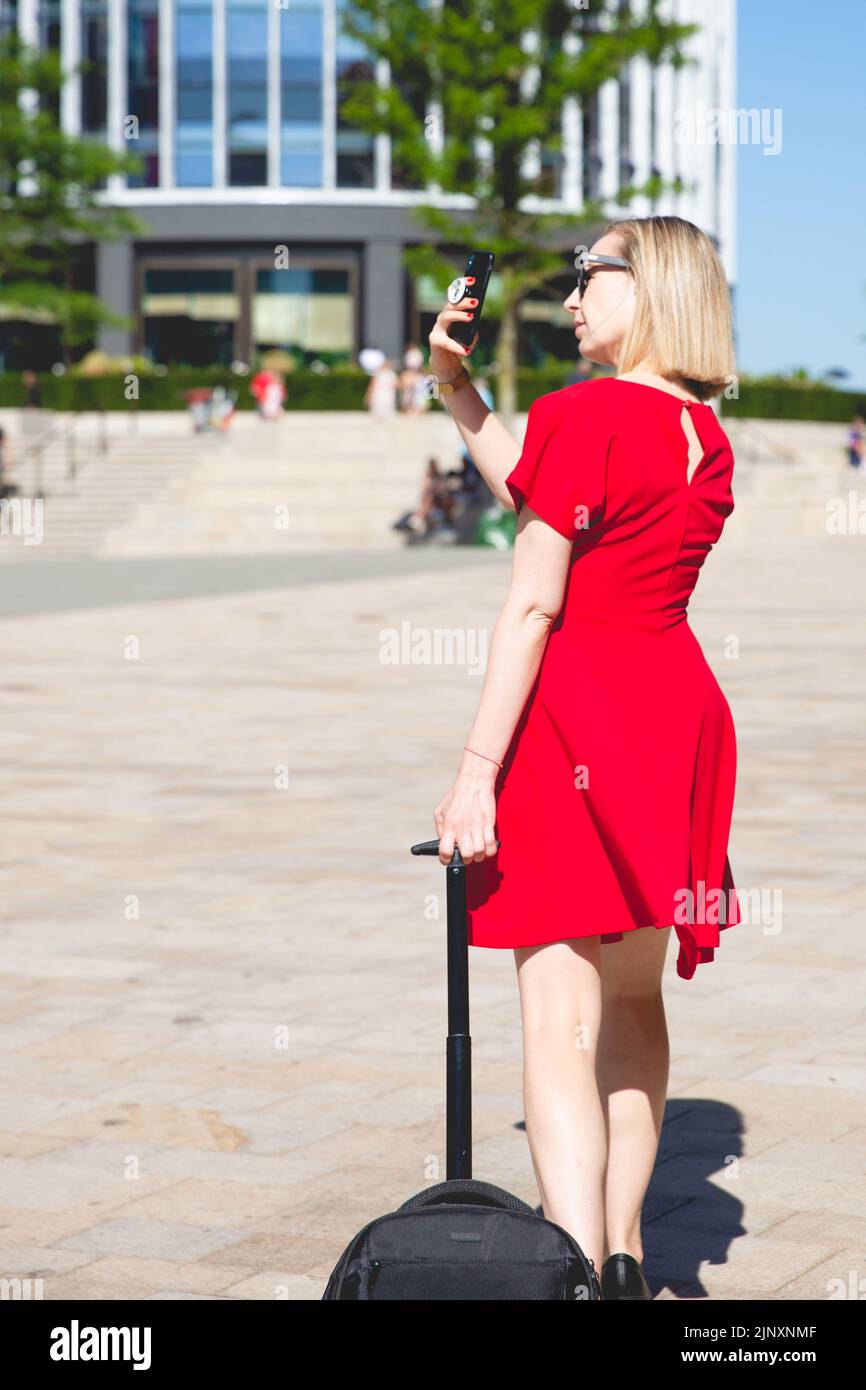 Rückansicht eines selbstbewussten jungen Mädchens in rotem Kleid, das mit ihrem Koffer durch eine Stadt läuft und ein Handy benutzt, um ein Foto zu machen Stockfoto
