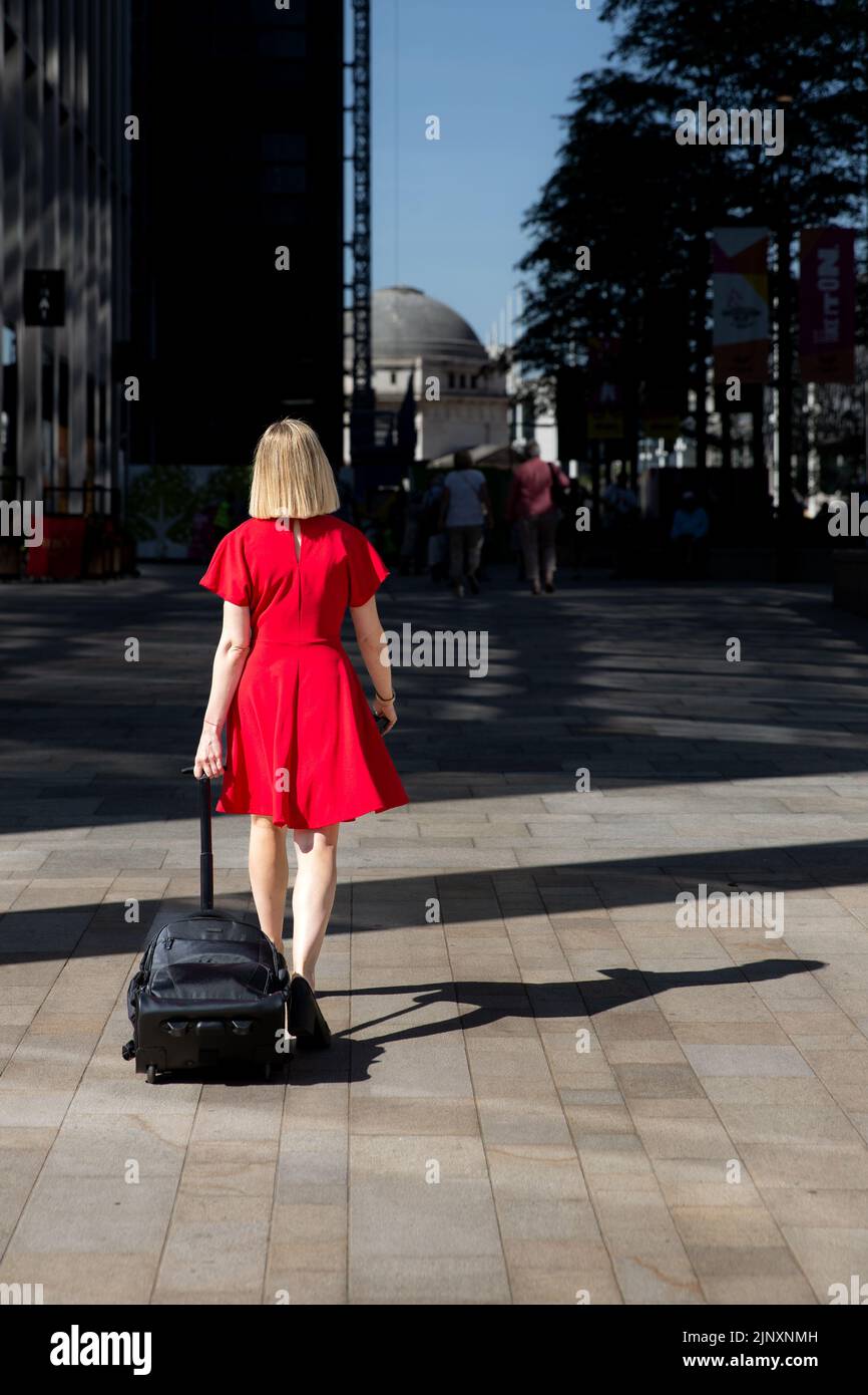 Rückansicht eines selbstbewussten jungen Mädchens in rotem Kleid, das mit ihrem Koffer durch eine Stadt geht und eine neue Reise durch das Leben beginnt Stockfoto