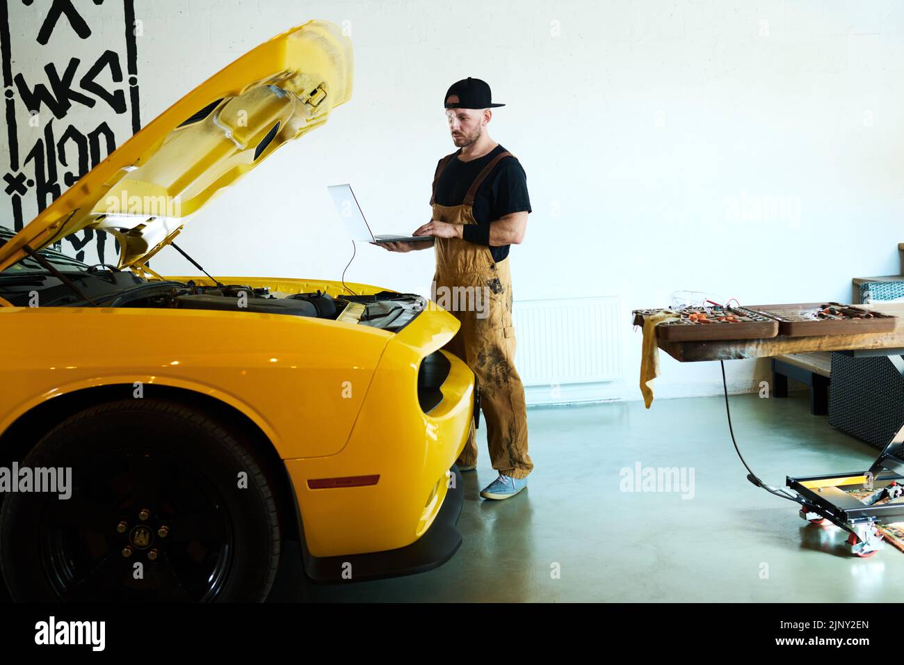 Junger Mechaniker in der Arbeitskleidung, der Laptop während der technischen Diagnose des Motors und anderer Details des Elektroautos der gelben Farbe hält Stockfoto
