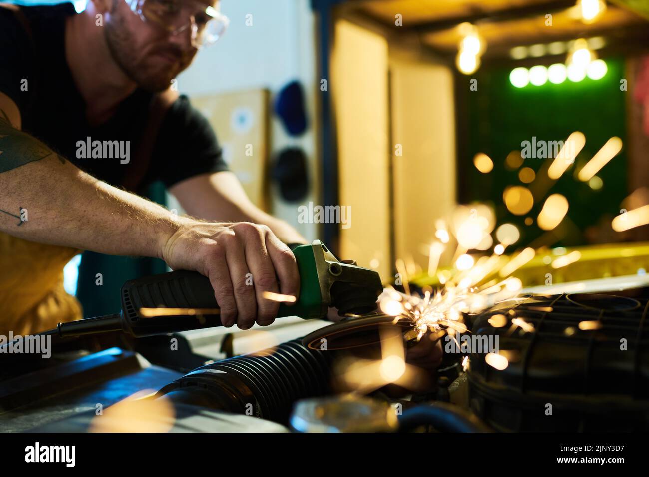 Junger Wartungsmitarbeiter mit elektrischem Handwerkzeug schleifend Details des Automotors beim Biegen über offene Haube während der Arbeit Stockfoto