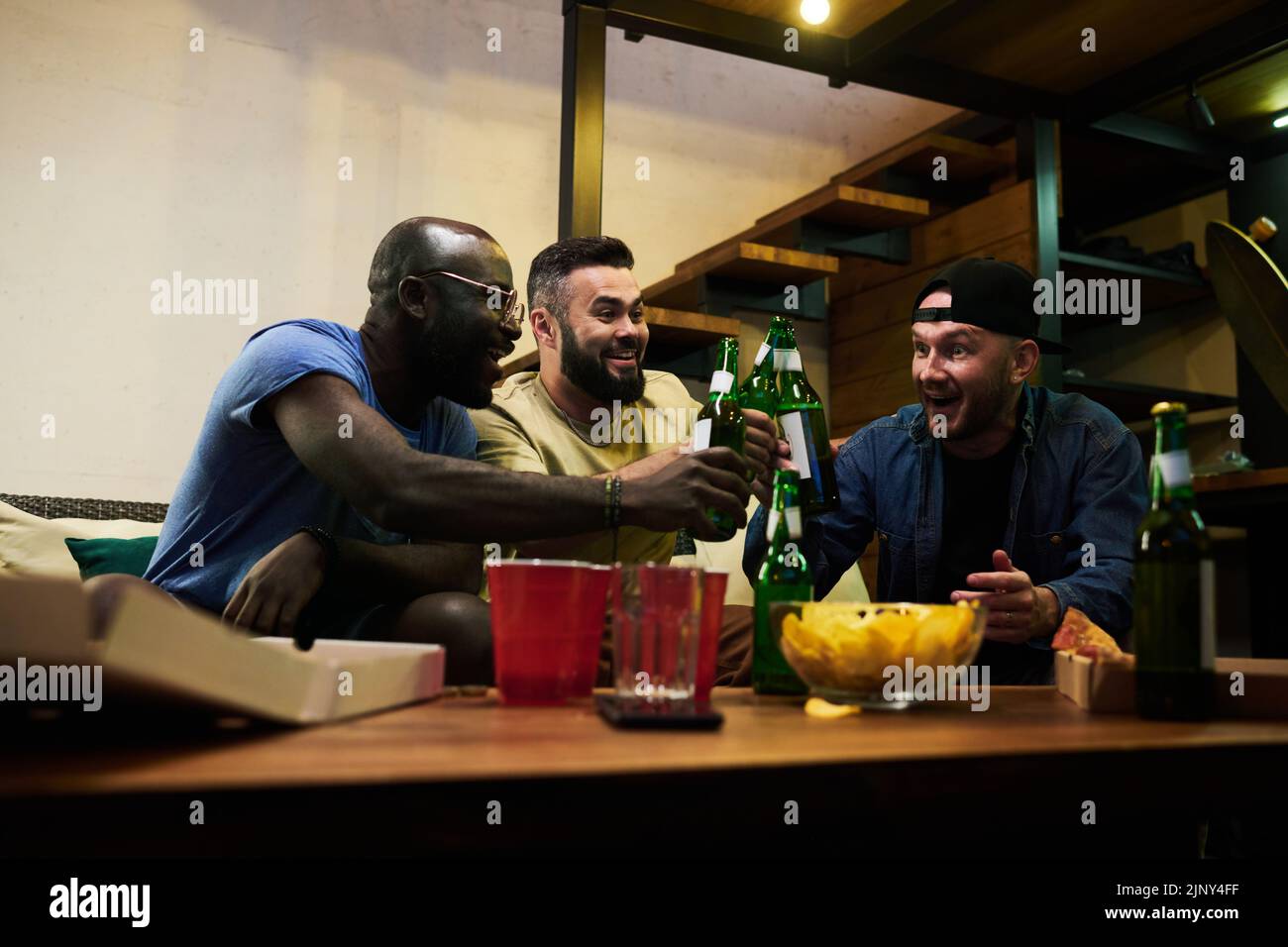 Junge, fröhliche, interracial Freunde und Fußballfans, die mit Bierflaschen über den Tisch mit Tassen und Snack klimmerten, während sie den Sieg feierten Stockfoto