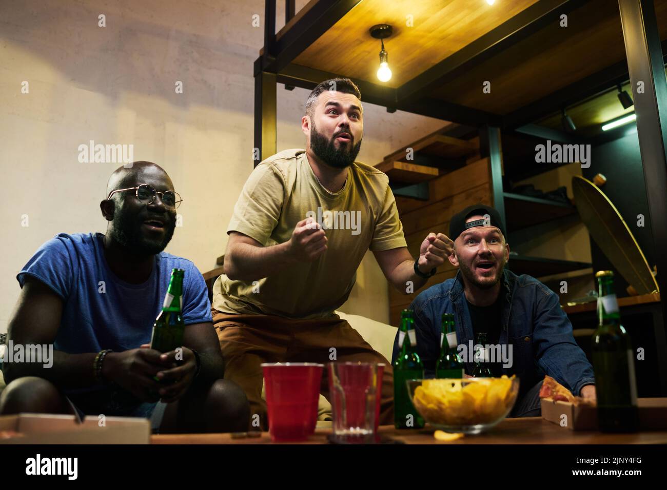 Drei junge multikulturelle Freunde, die ihre Freude über Fußballspiele zum Ausdruck bringen, während sie in der Garage im fernsehen ausgestrahlt werden Stockfoto