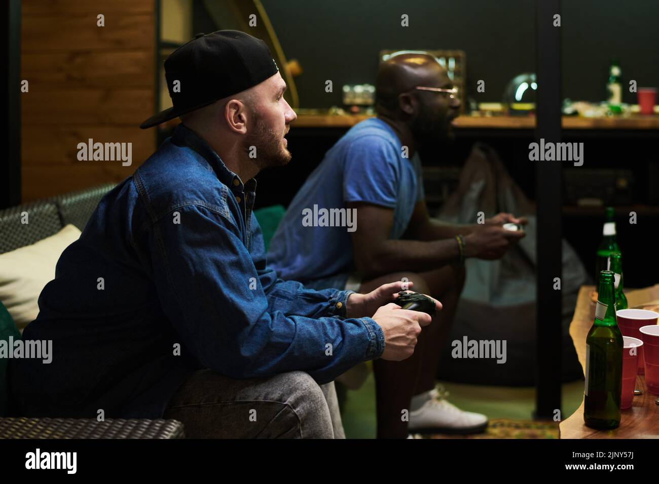 Angespannter Mann in Casualwear, der Tasten auf dem Joystick drückt, während er mit seinem Kumpel ein Videospiel spielt, während beide mit Bier vor dem Tisch sitzen Stockfoto