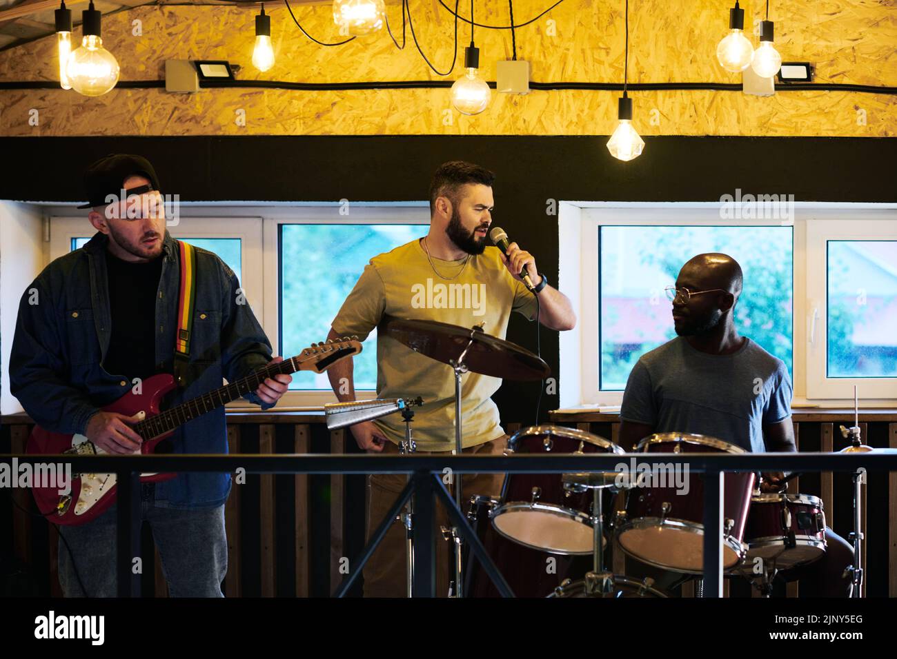 Porträt von drei jungen interkulturellen Männern in der Casualwear, die im Mikrofon singen und während der Wiederholung Musikinstrumente spielen Stockfoto