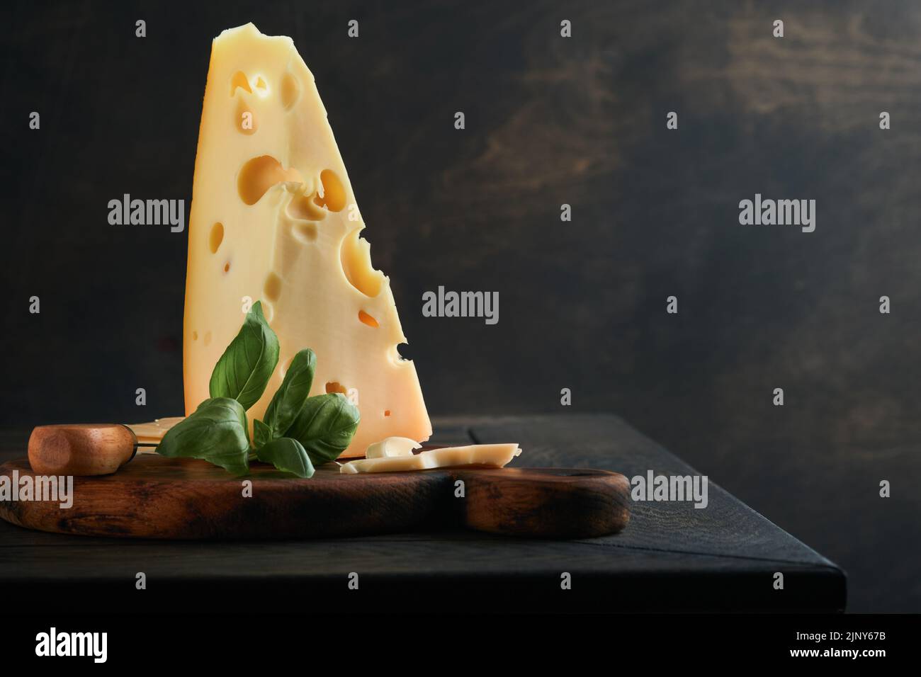 Käse. Stück Käsedreieck, Schweizer Käse mit Basilikum und ein Messer auf einem Servierbrett auf dunklem Holztischhintergrund. Käsehintergrund. Stockfoto