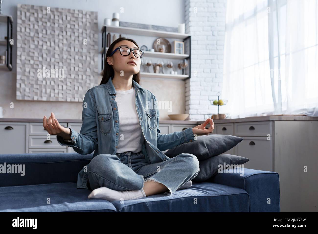 Schöne asiatische Frau, die sich zu Hause entspannt, auf dem Sofa sitzt und in Lotuspose meditiert, Atemübungen im Wohnzimmer macht Stockfoto