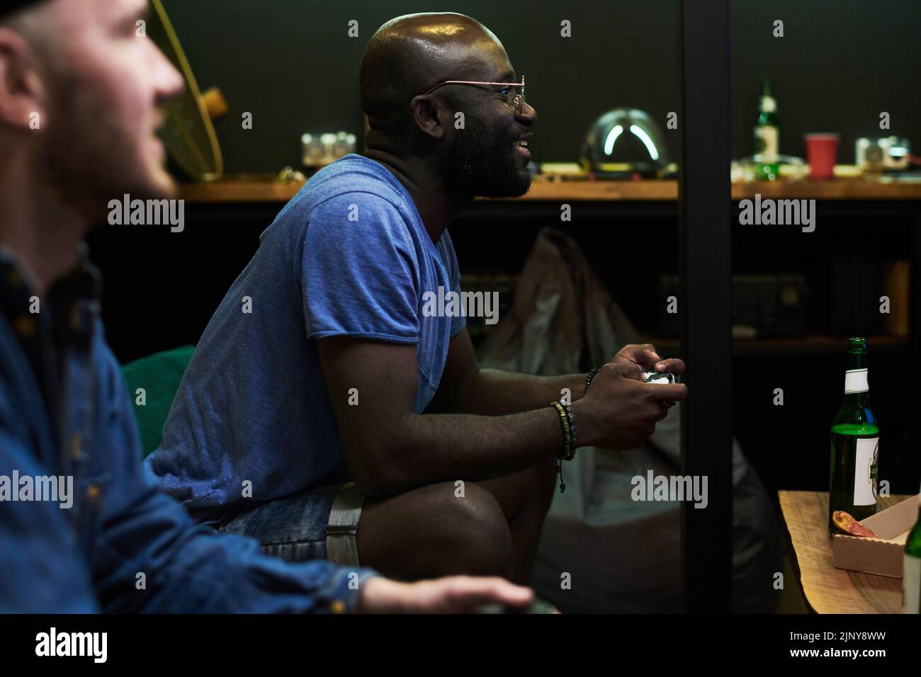Glücklicher junger afroamerikanischer Mann in blauem T-Shirt, der Videospiel spielt, während er auf der Couch sitzt und Tasten auf dem Gamepad drückt Stockfoto