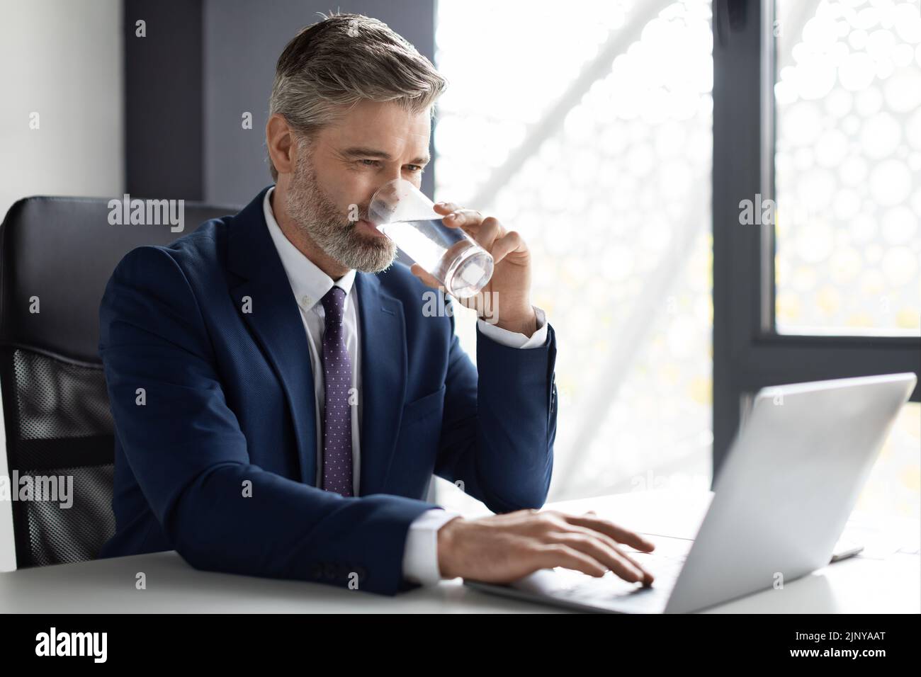 Geschäftsmann Mittleren Alters, Der Während Der Arbeit Mit Einem Laptop Im Büro Wasser Trinkt Stockfoto