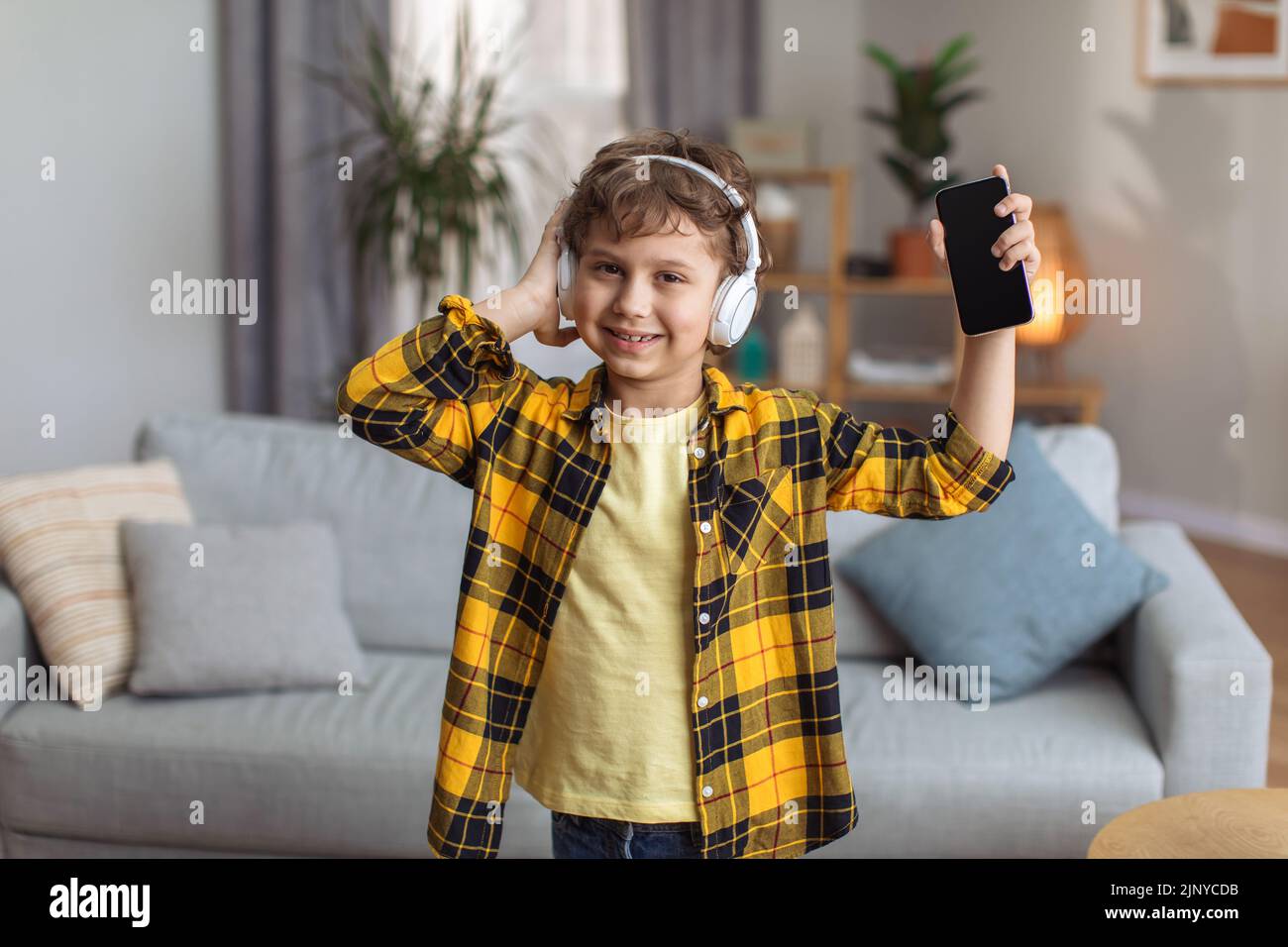Musik-App für Kinder. Fröhlicher kleiner Junge, der Musik über Kopfhörer genießt und der Kamera das Smartphone mit leerem Bildschirm zeigt Stockfoto