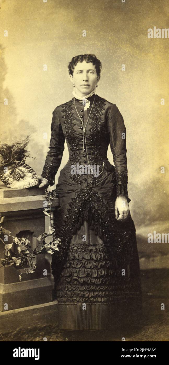 Ende 19. Century Photo Studio Porträt einer formellen ernsthaften Frau, die in stilvoller, zeitlicher Kleidung mit Hintergrund um 1870 - 1880s steht Stockfoto